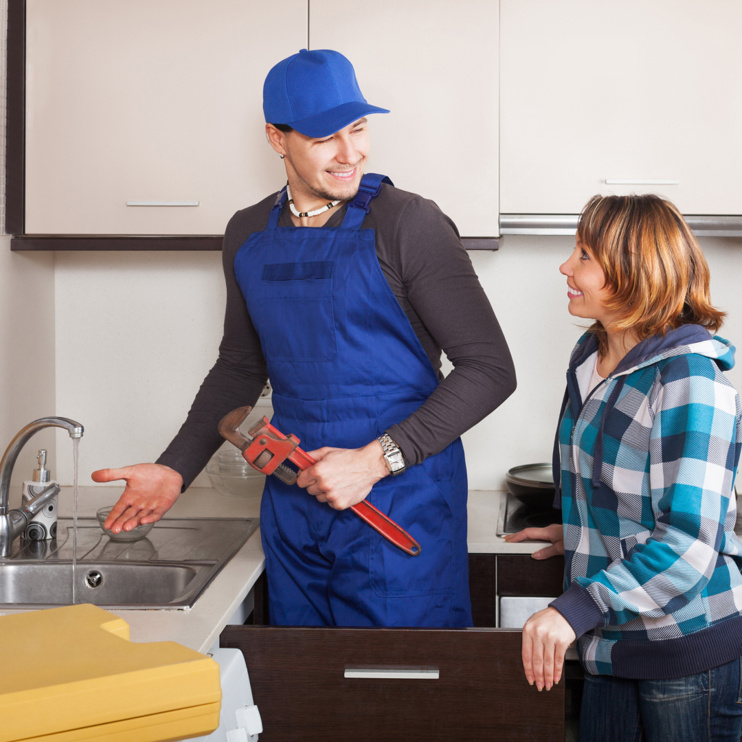 A plumber in a blue uniform showcases his Drain Magic skills, holding a wrench and chatting with a woman in her Pennsylvania kitchen near the sink.